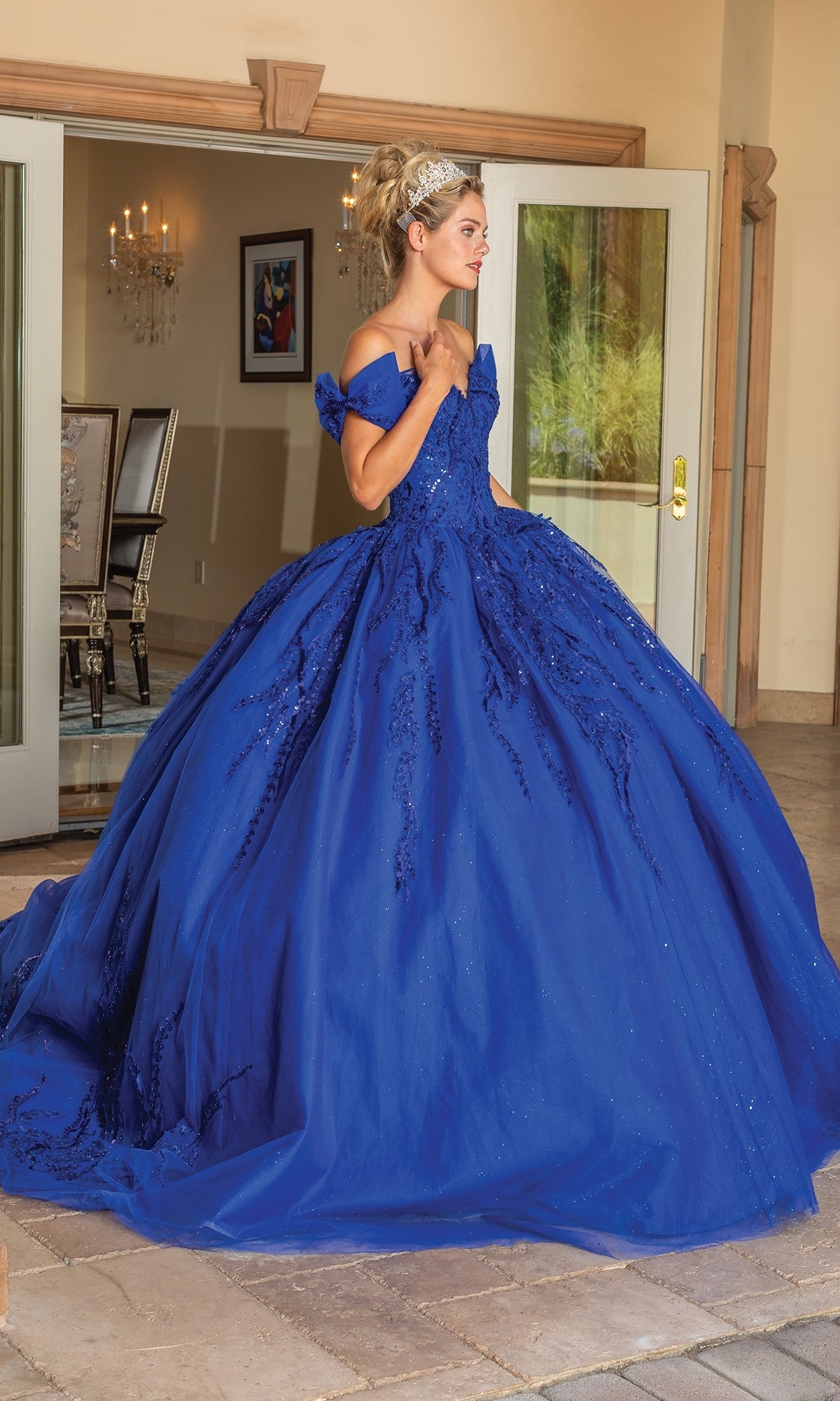 A woman wearing the Quinceanera Dress 1780 By Dancing Queen, a royal blue off-the-shoulder ball gown, stands by an open doorway and looks to the side.